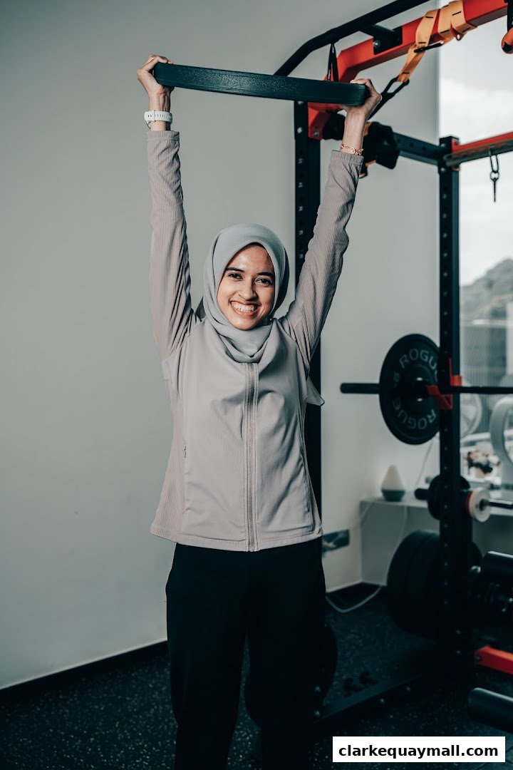 A woman in athletic wear and a hijab smiles while holding onto a pull-up bar in a gym.