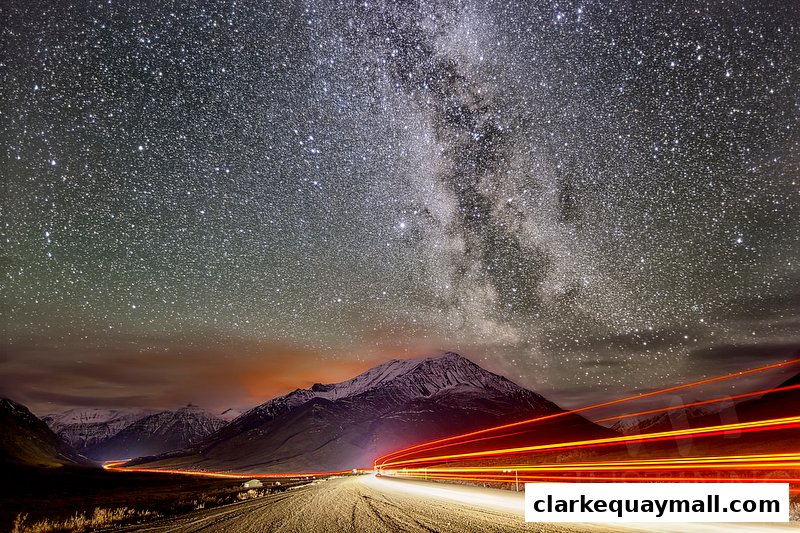 A stunning night landscape with the Milky Way, snow-capped mountains, and light trails from a road.