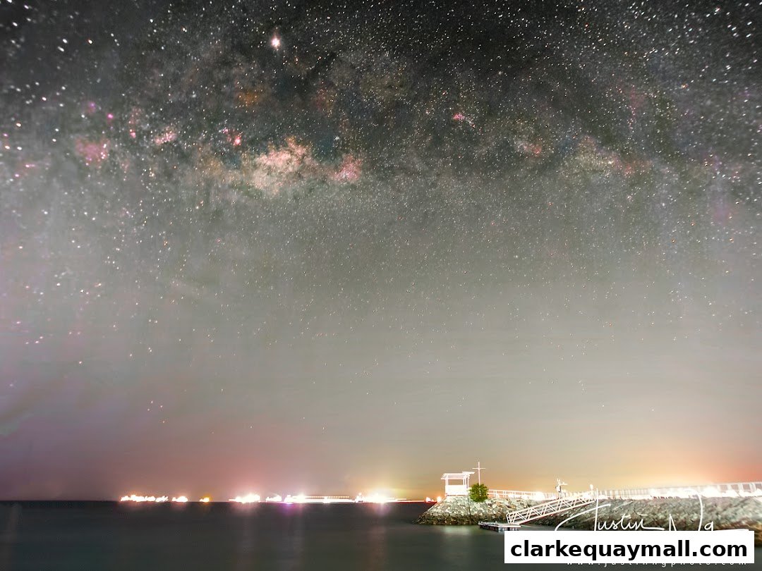 The Milky Way galaxy stretches across the night sky above a calm sea and a pier.