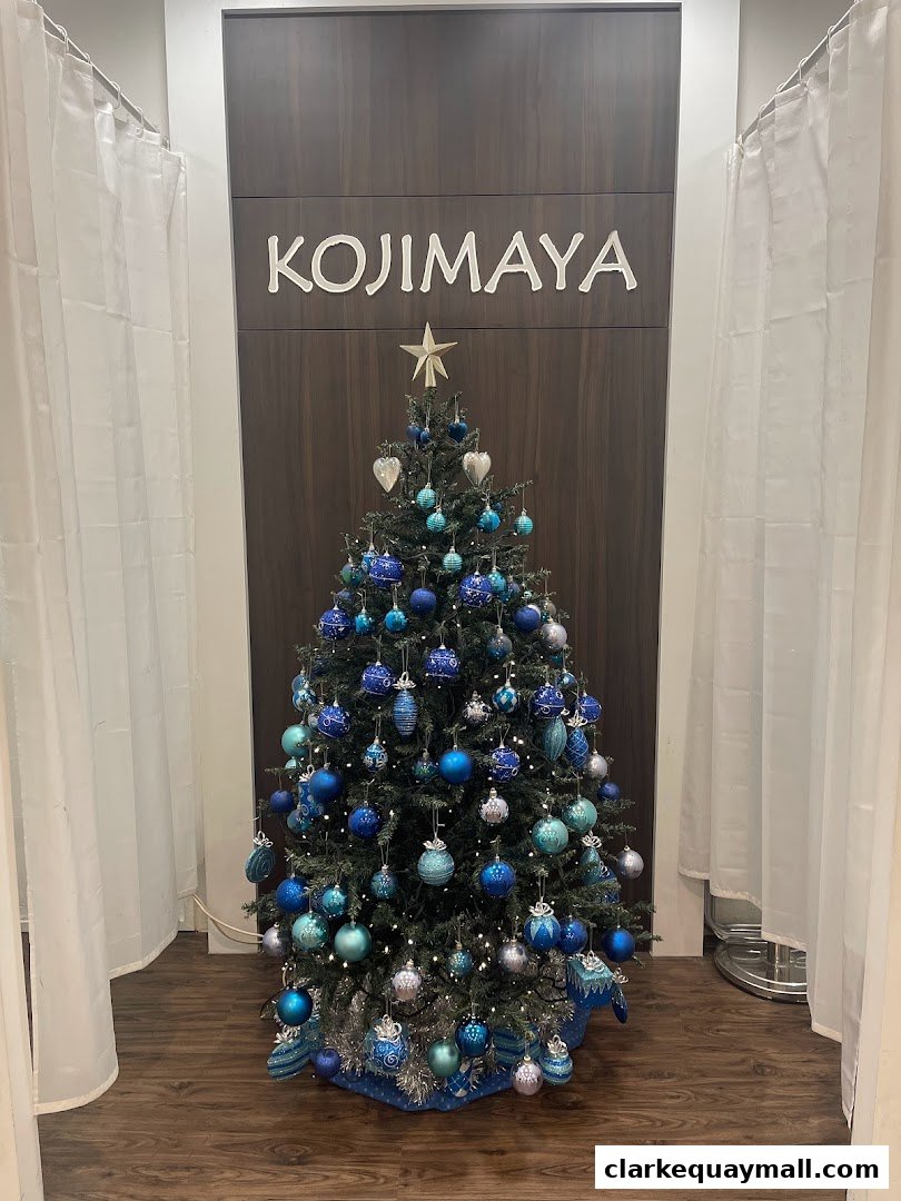 A decorated Christmas tree with blue and silver ornaments in front of the KOJIMAYA logo.