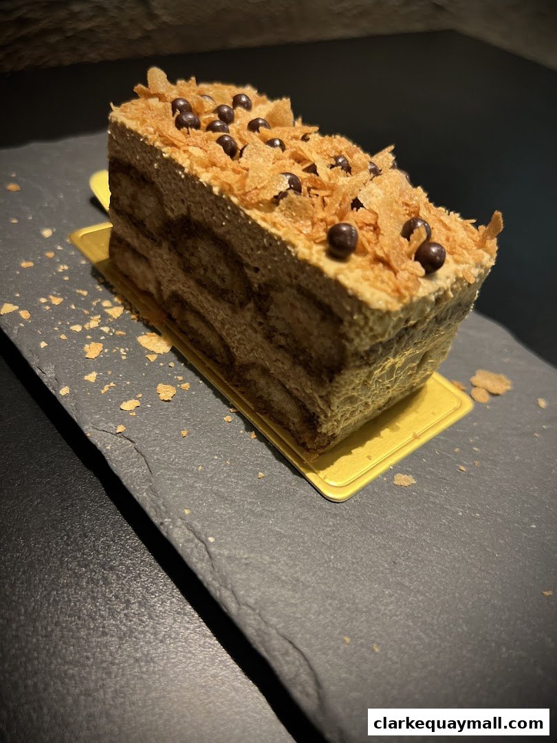 A slice of tiramisu cake with chocolate balls and crispy flakes on top.