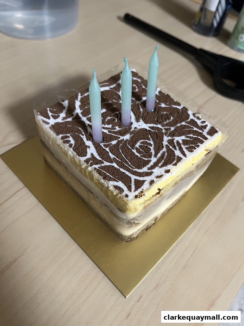 A square tiramisu cake with three pastel candles on a golden cake board.
