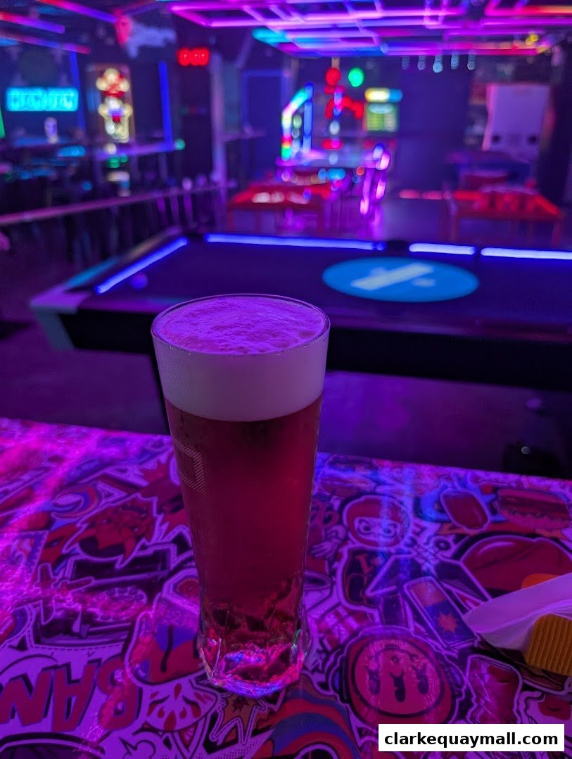 A tall glass of pinkish beverage with foam sits on a colorful, sticker-covered table in a neon-lit arcade bar.