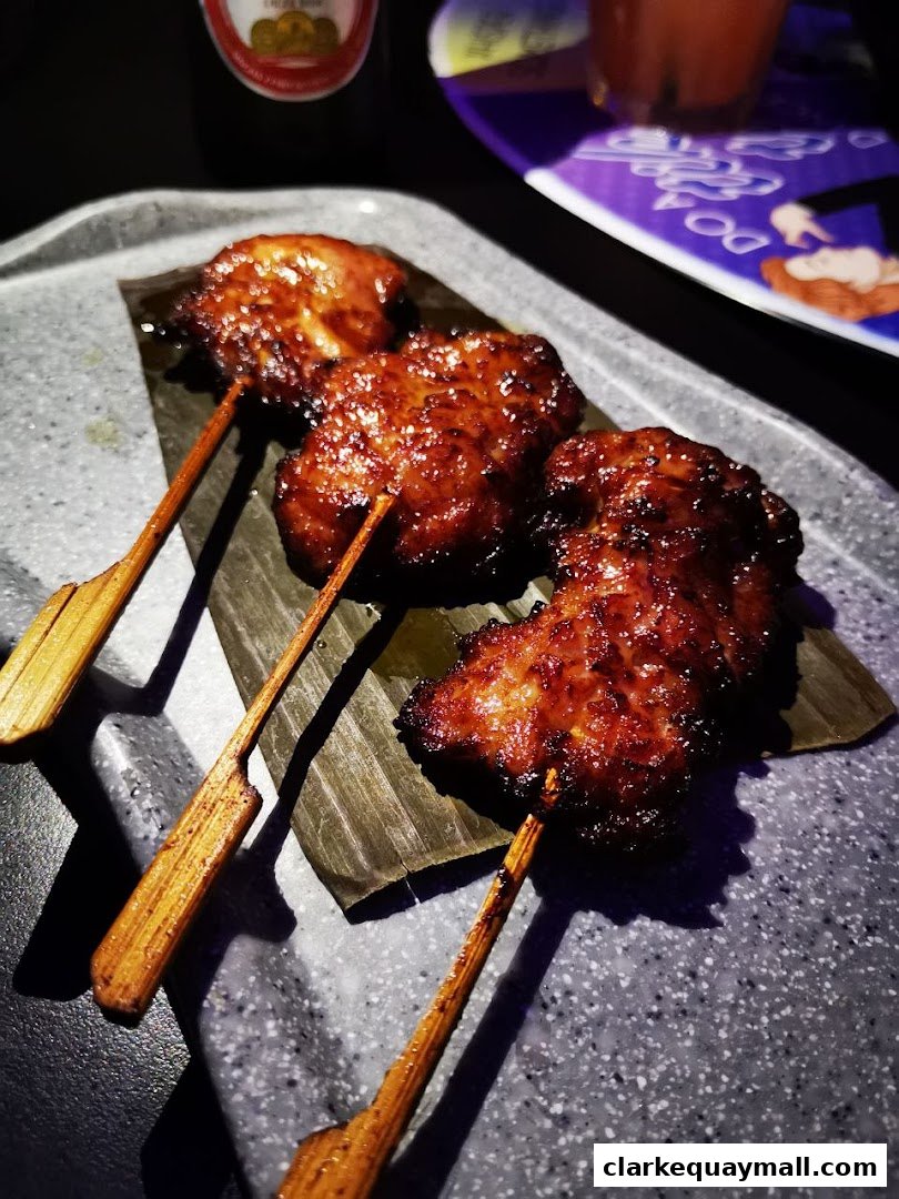 Three glazed skewers of meat are presented on a textured gray plate with banana leaves.