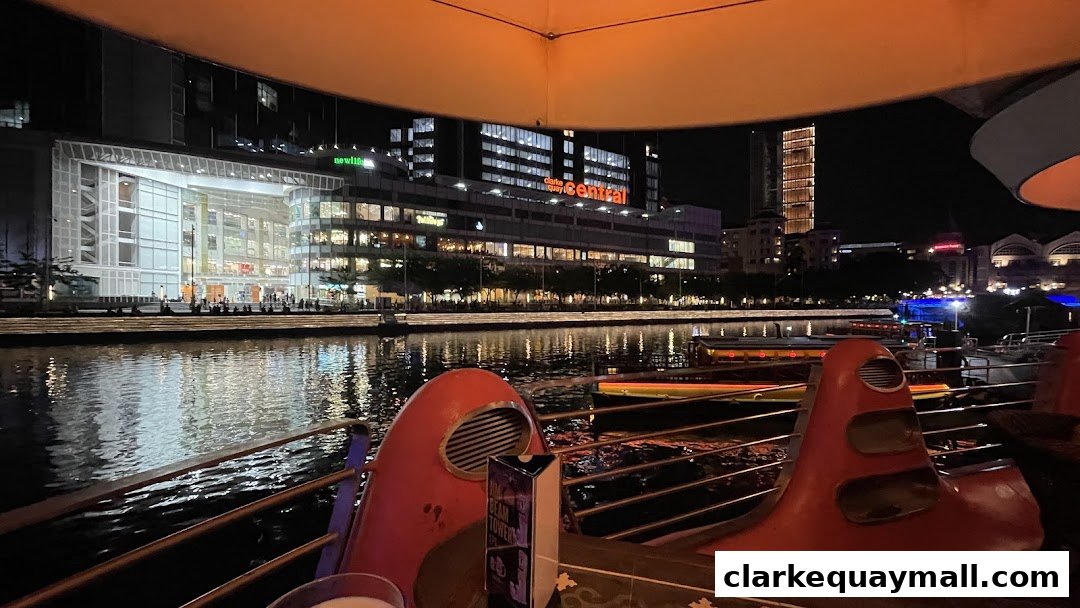 Night view of a riverfront with illuminated buildings and boats, seen from a seating area.