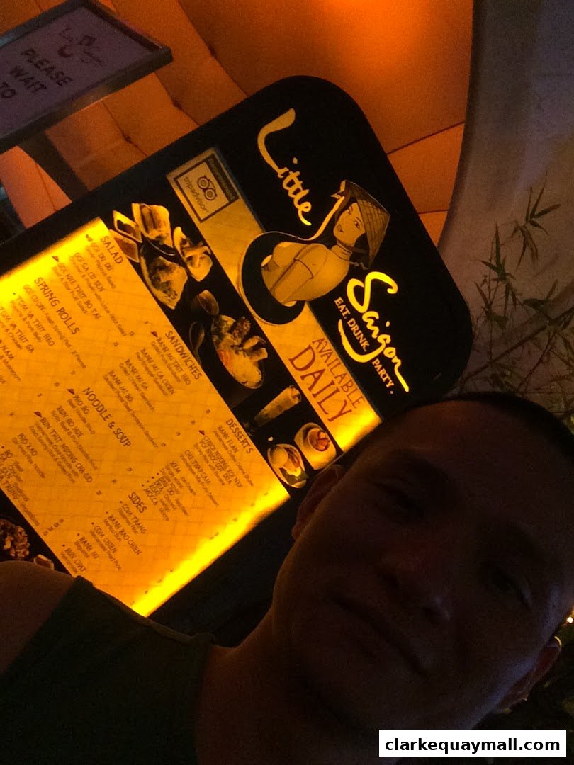 A person takes a selfie in front of a brightly lit menu for Little Saigon restaurant.