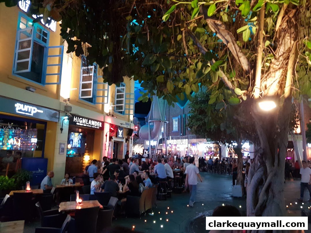 A lively outdoor dining area at night with people seated at tables under trees and illuminated shops.