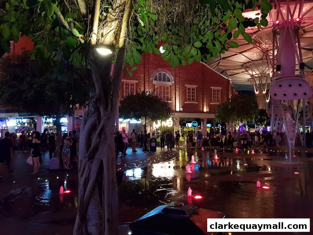 A lively evening scene with people enjoying a water fountain display in front of brick buildings.