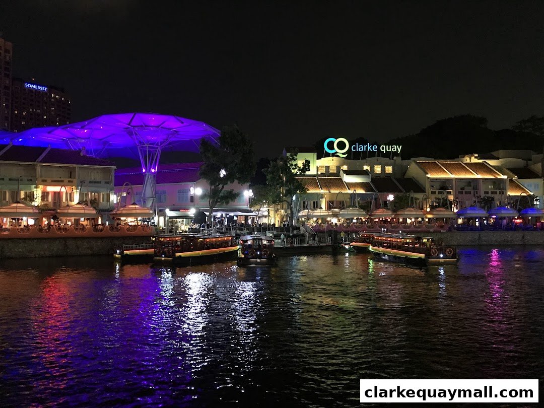 Night view of Clarke Quay with illuminated buildings, boats on the river, and a purple canopy.