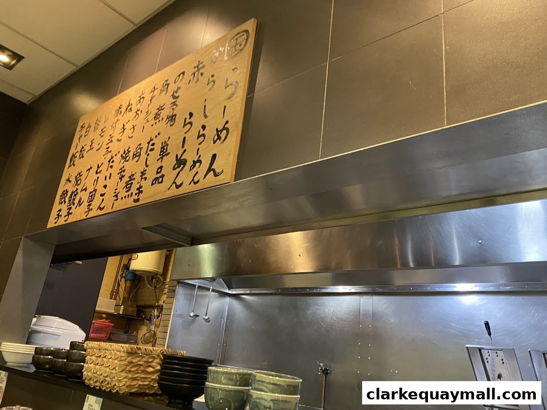 A wooden board with Japanese calligraphy menu items hangs above a stainless steel kitchen counter.