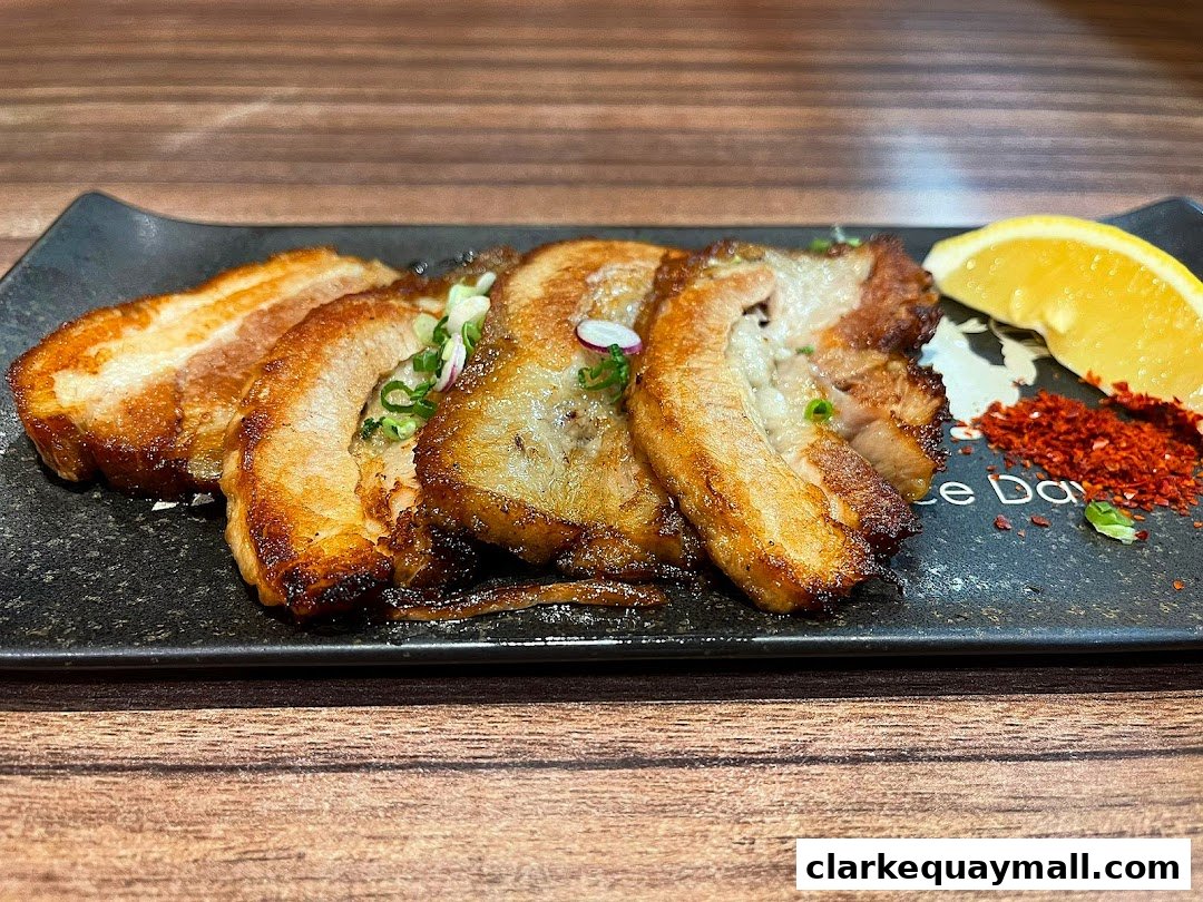 A close-up of tender, grilled pork belly slices served with a lemon wedge and chili flakes.
