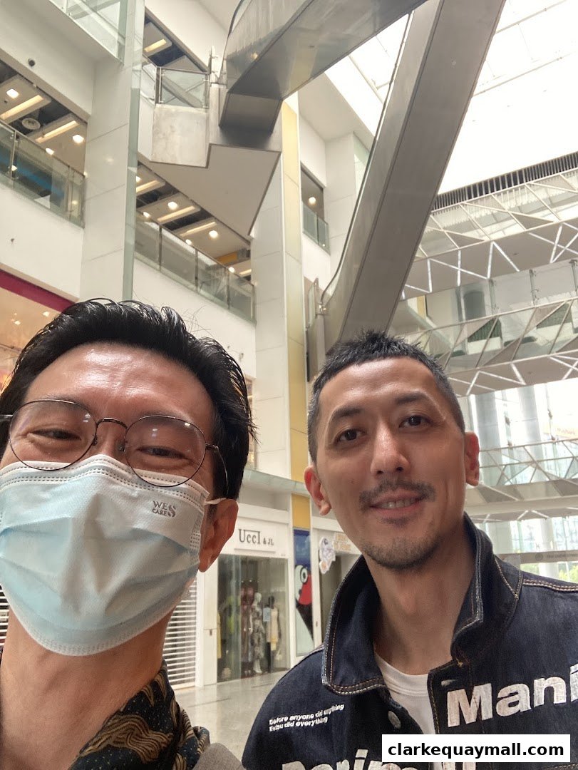 Two men pose for a selfie in a modern shopping mall with escalators and shops visible.