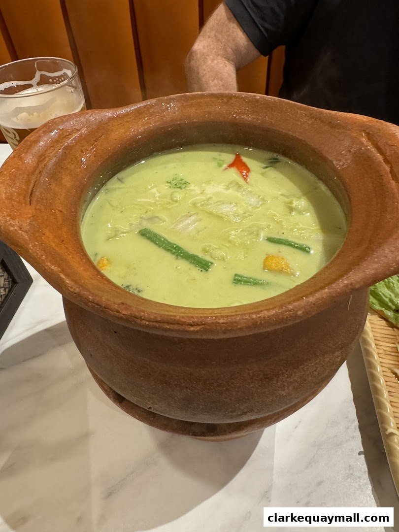 A close-up of a steaming bowl of green curry with vegetables and a glass of beer.