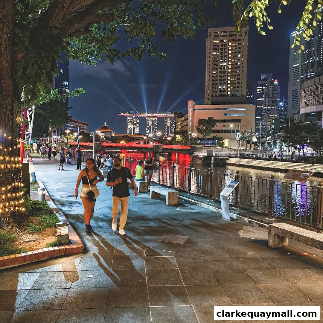 A couple walks along a riverfront promenade at night with city skyline and illuminated buildings in the background.