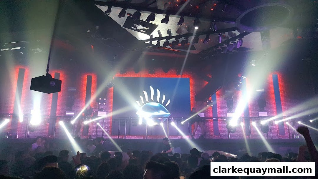 A crowded nightclub with a DJ on stage, bright lights, and a large screen displaying a logo.