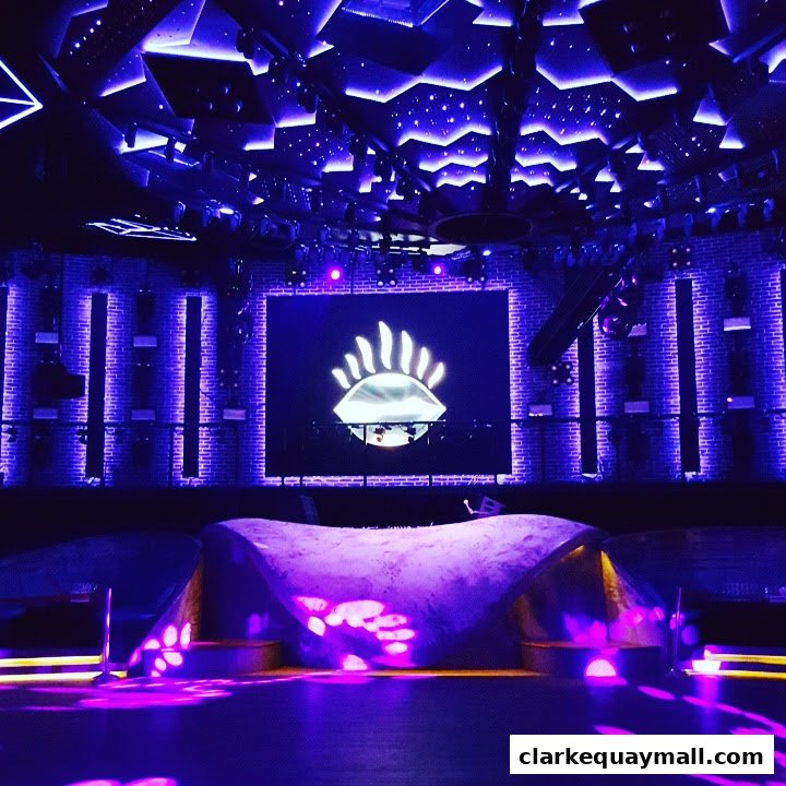 An interior shot of a club with a stage, a large screen displaying a logo, and purple lighting.