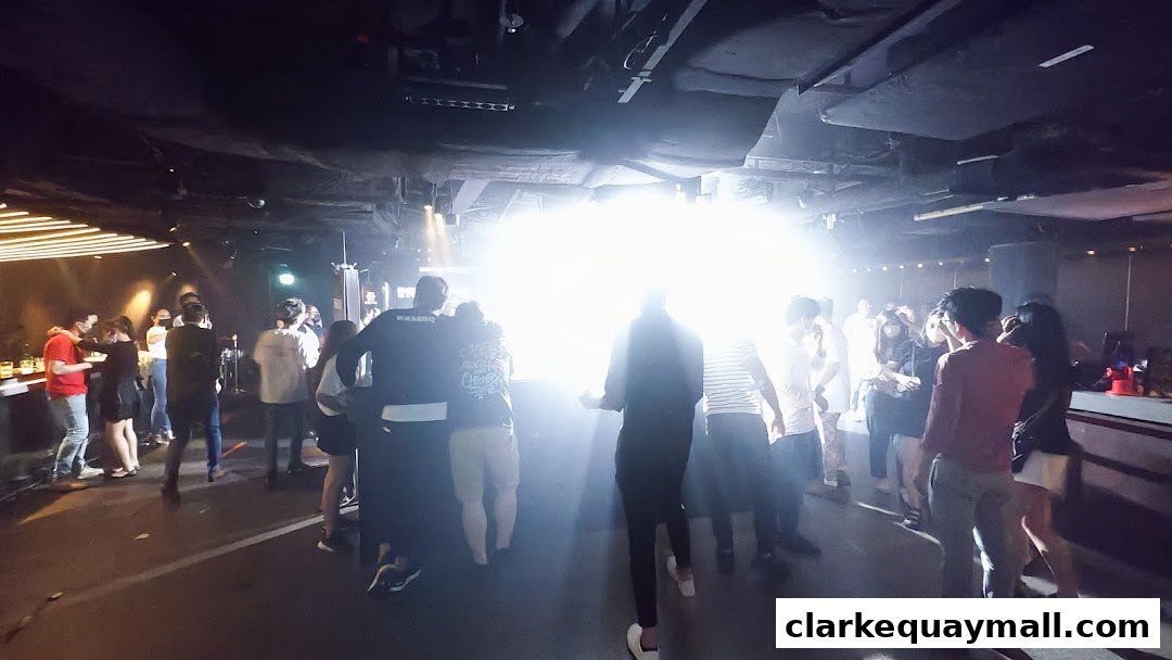 People gather inside a dimly lit venue with bright lights and a DJ booth.