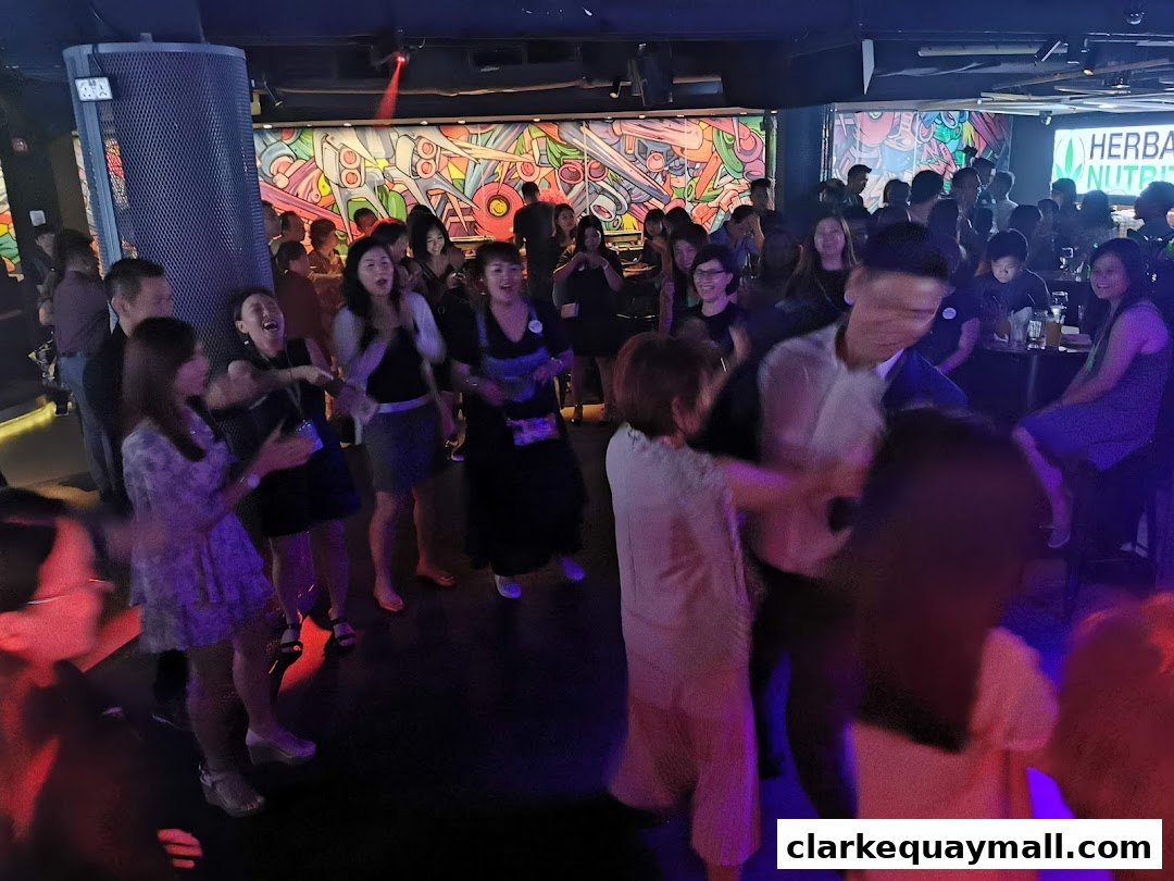 People dancing and socializing in a dimly lit venue with colorful wall art and a prominent Herbalife Nutrition logo.