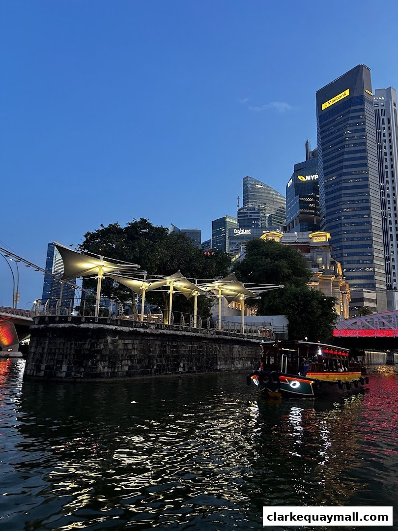 A river cruise boat sails past a waterfront seating area with city skyscrapers in the background at dusk.