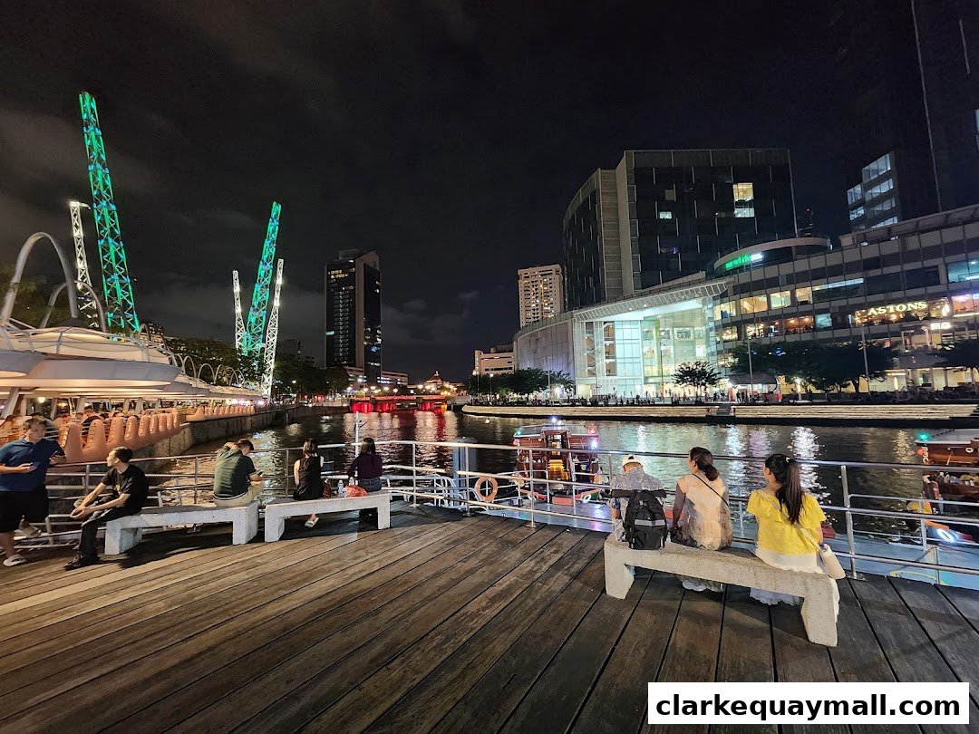 People relax on benches by a river at night, with illuminated buildings and structures in the background.