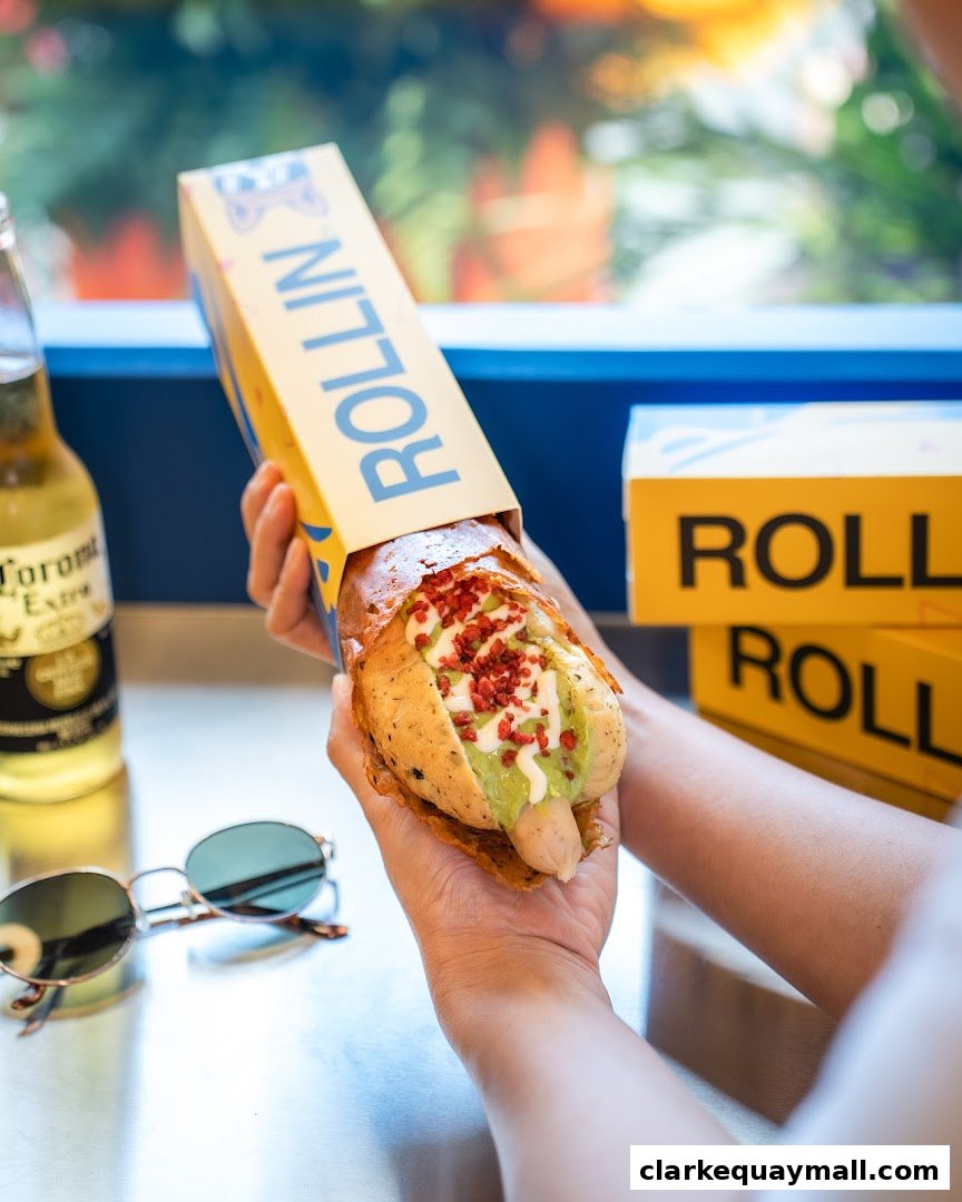 A person holds a gourmet hot dog topped with avocado and bacon bits in a branded box.