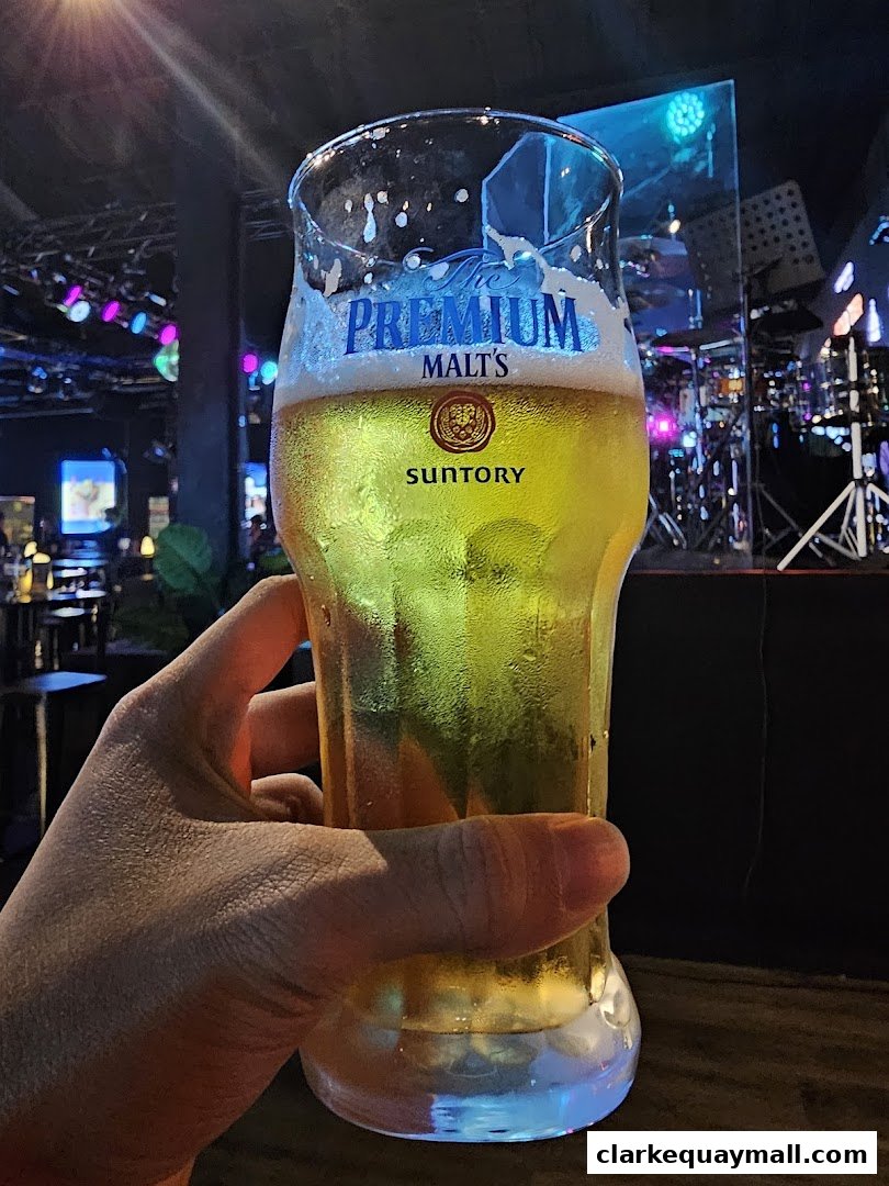 A hand holds a frosty glass of premium malt beer in a dimly lit venue with a stage.
