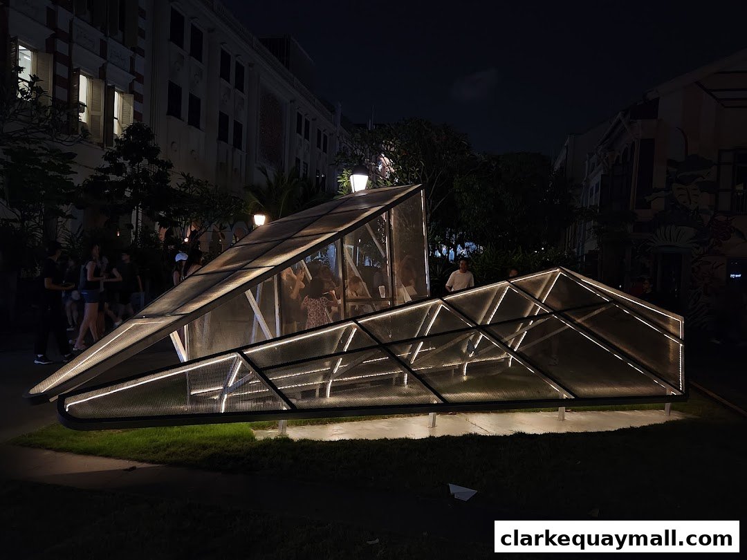 A modern, illuminated glass structure with people inside, set against a nighttime cityscape.