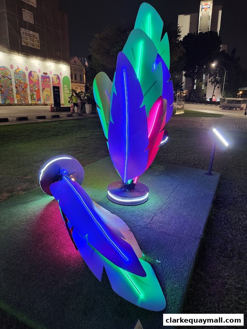 Colorful feather-shaped light installations illuminate a grassy area at night.