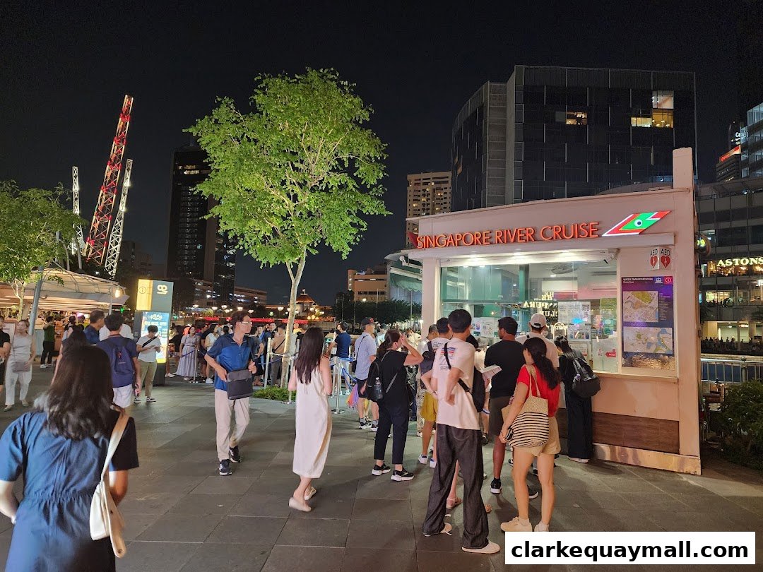 People gather at the Singapore River Cruise booth at night, with city lights and a Ferris wheel in the background.