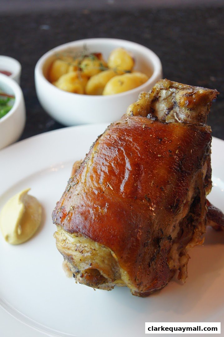 A close-up of a roasted pork knuckle with a side of potatoes and mustard.