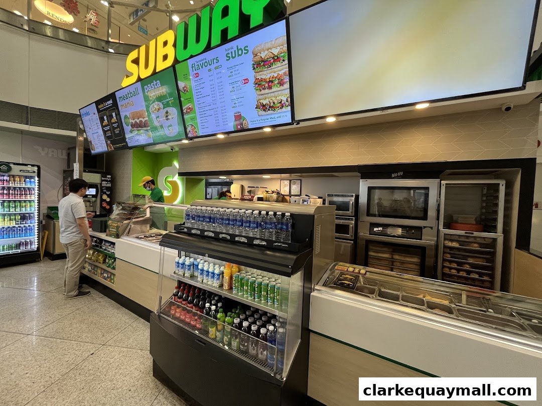 A Subway restaurant interior with a customer ordering at the counter and digital menus displaying food options.
