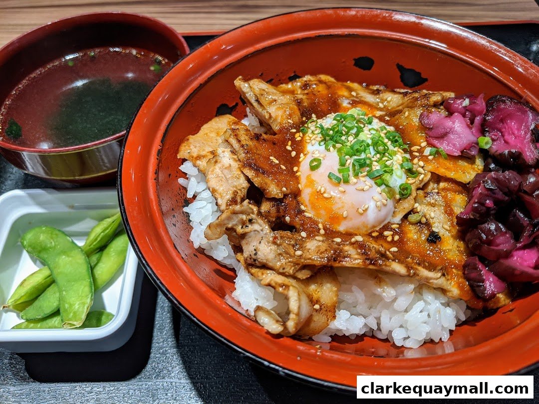 A delicious bowl of rice topped with grilled pork, a soft-boiled egg, and pickled vegetables, served with edamame and soup.