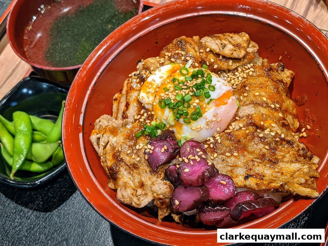 A delicious bowl of rice topped with grilled meat, a soft-boiled egg, and pickled vegetables, served with edamame and soup.