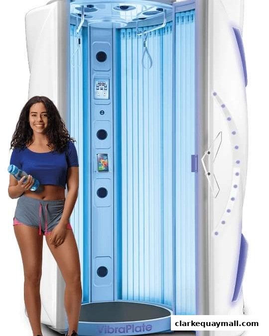 A smiling woman stands next to a modern, illuminated tanning booth.