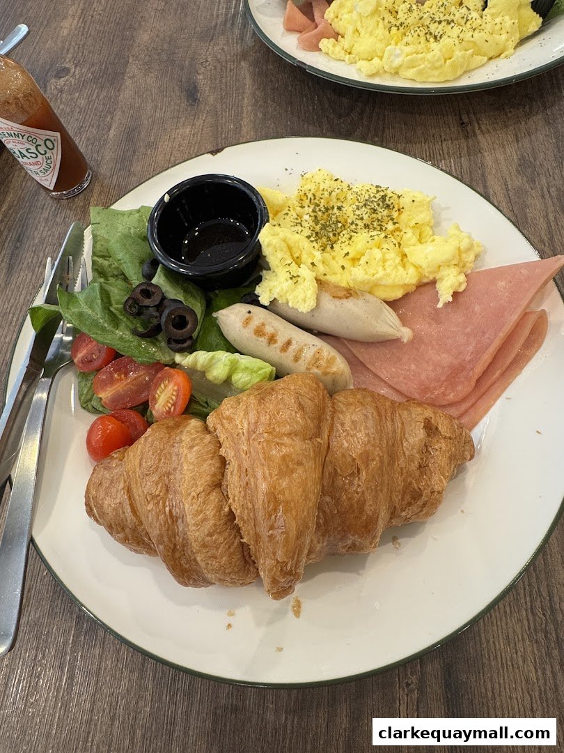 A delicious breakfast spread featuring a croissant, scrambled eggs, sausages, ham, salad, and a bottle of Tabasco sauce.