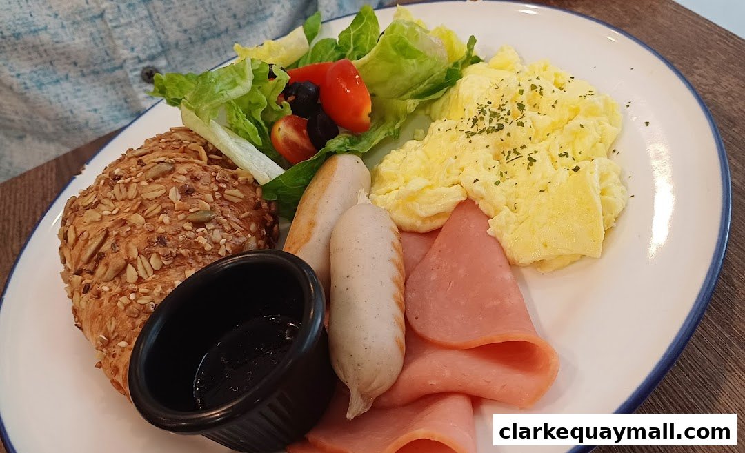 A hearty breakfast plate with scrambled eggs, sausages, ham, a seeded bun, and a small salad.