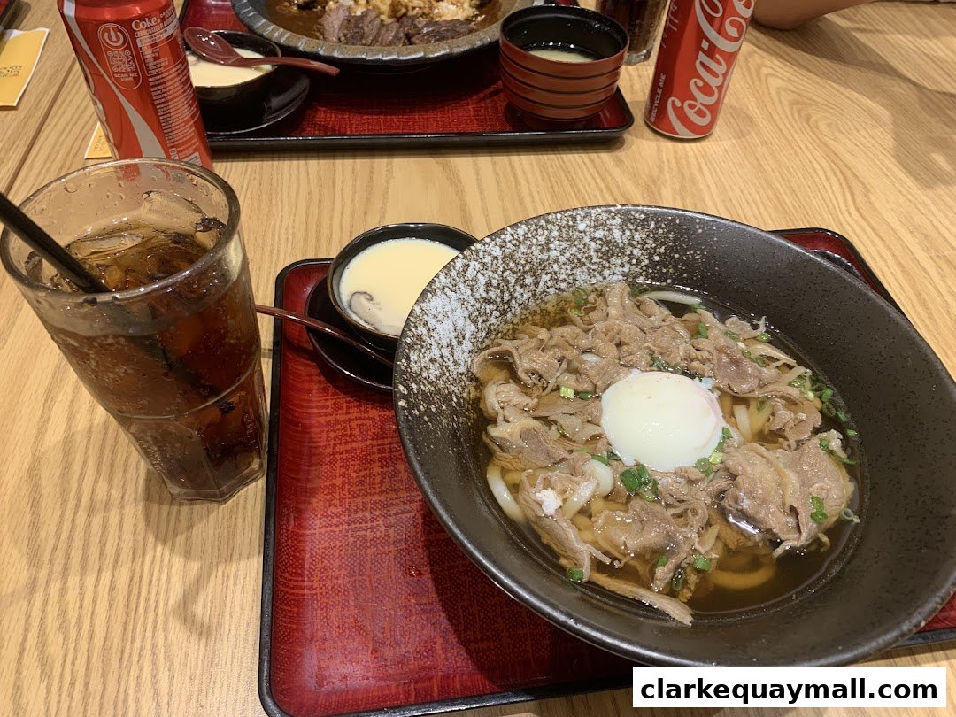 A close-up of a delicious beef udon dish with a soft-boiled egg and a refreshing cola.