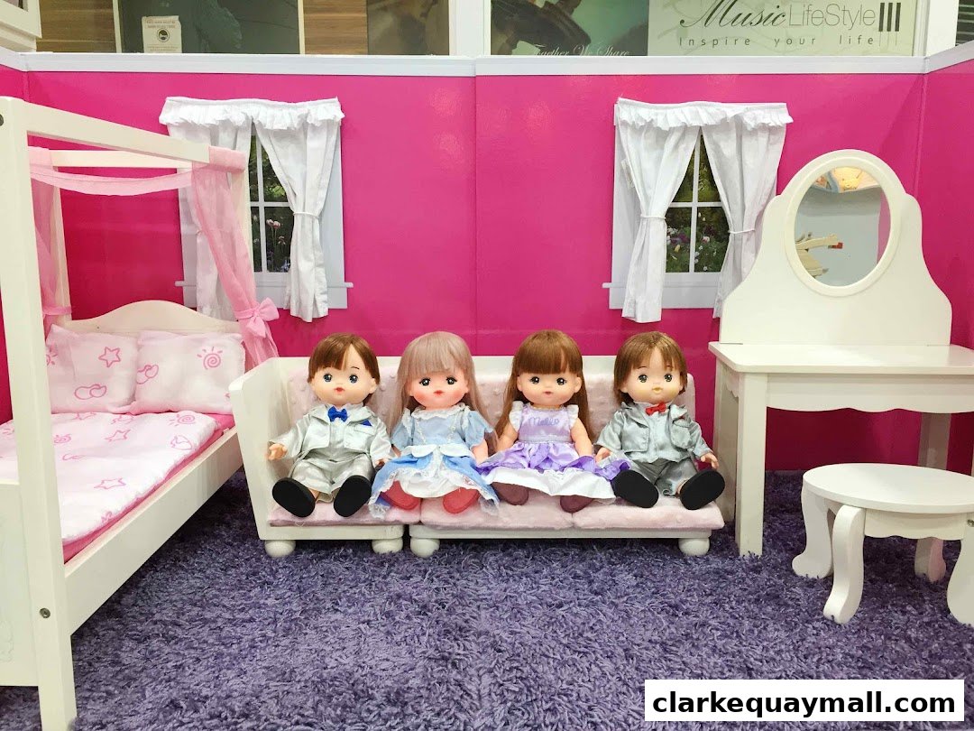 Four dolls are sitting on a pink couch in a room with a doll bed and vanity.