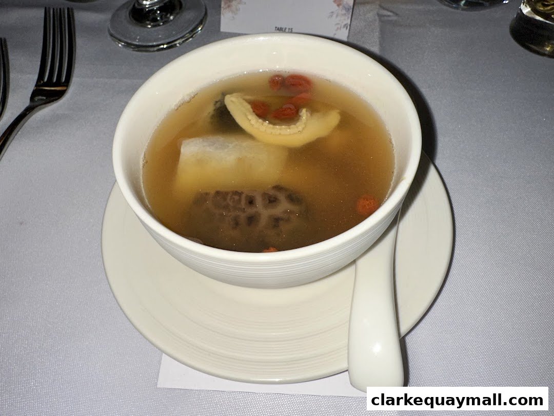 A bowl of clear soup with various ingredients and a spoon on a white saucer.