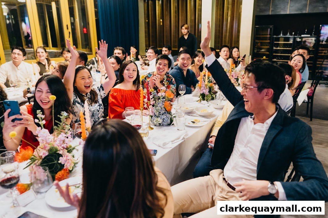 A group of people are seated at a long table, enjoying a meal and raising their hands in excitement.