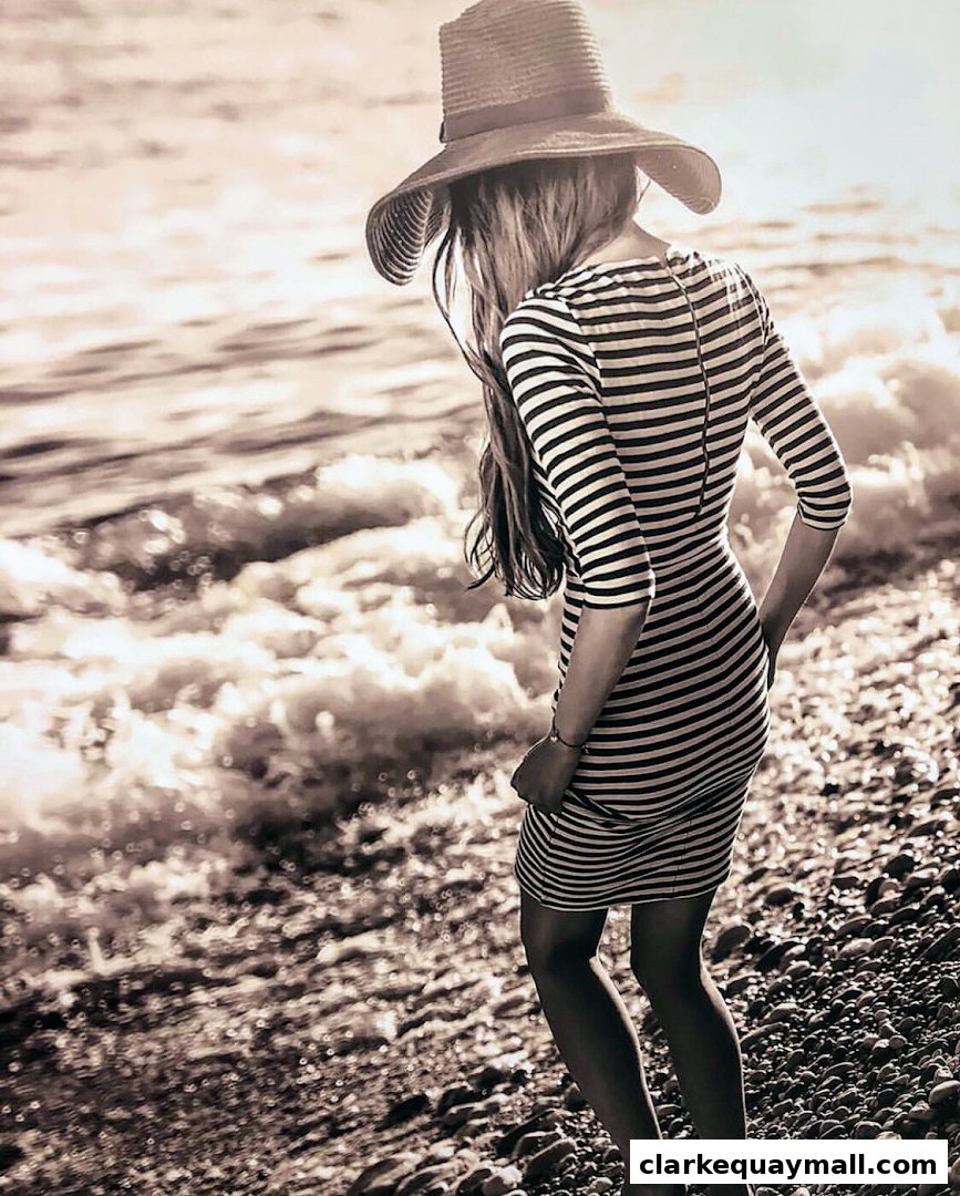 A woman in a striped dress and hat stands on a pebble beach by the sea.