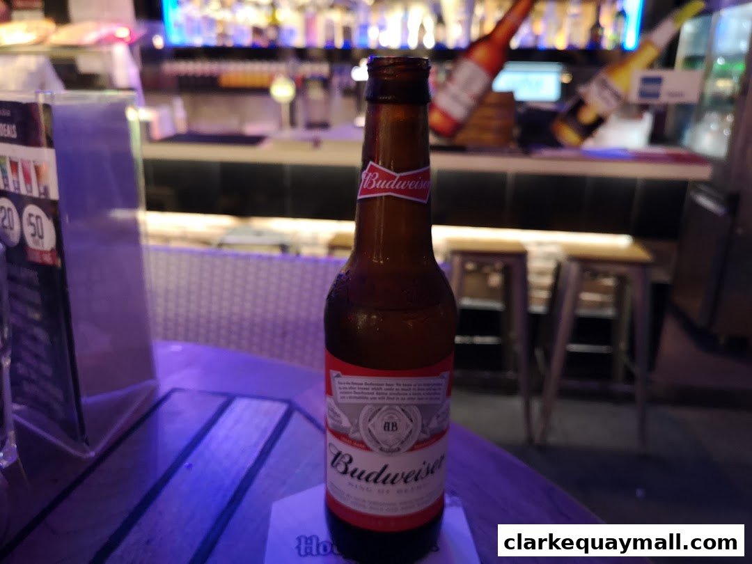 A Budweiser beer bottle sits on a table in front of a bar with stools and bottles.