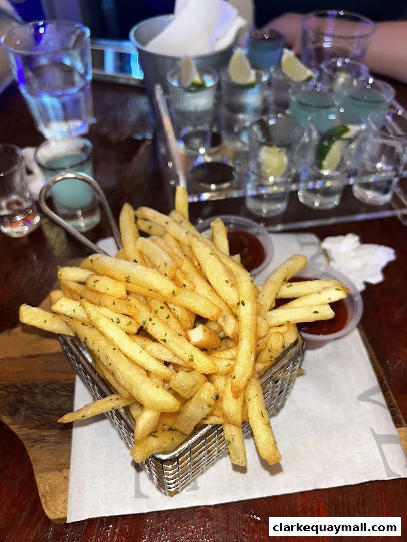 A basket of golden french fries with dipping sauce and a tray of colorful shots in the background.