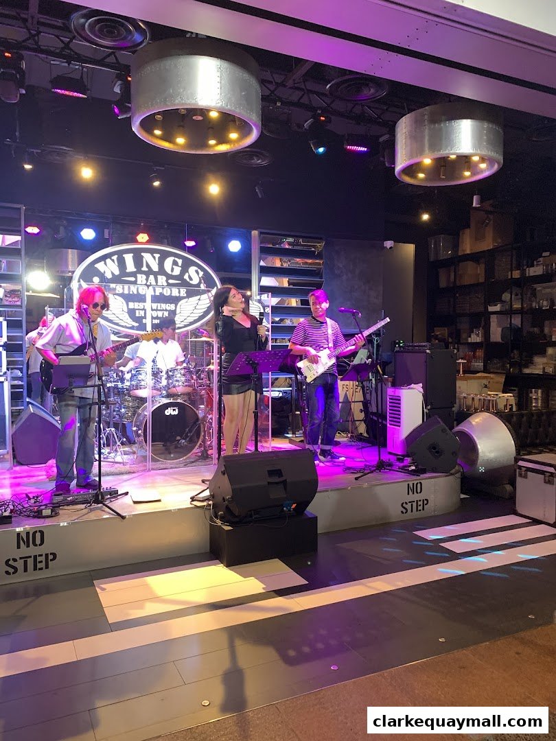 A live band performs on stage at Wings Bar & Grill, Singapore, with a singer and musicians playing instruments.