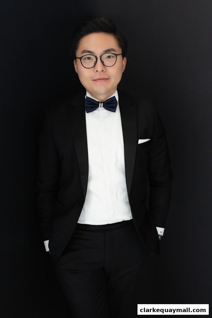 A man in a tuxedo with a bow tie and glasses poses against a black background.