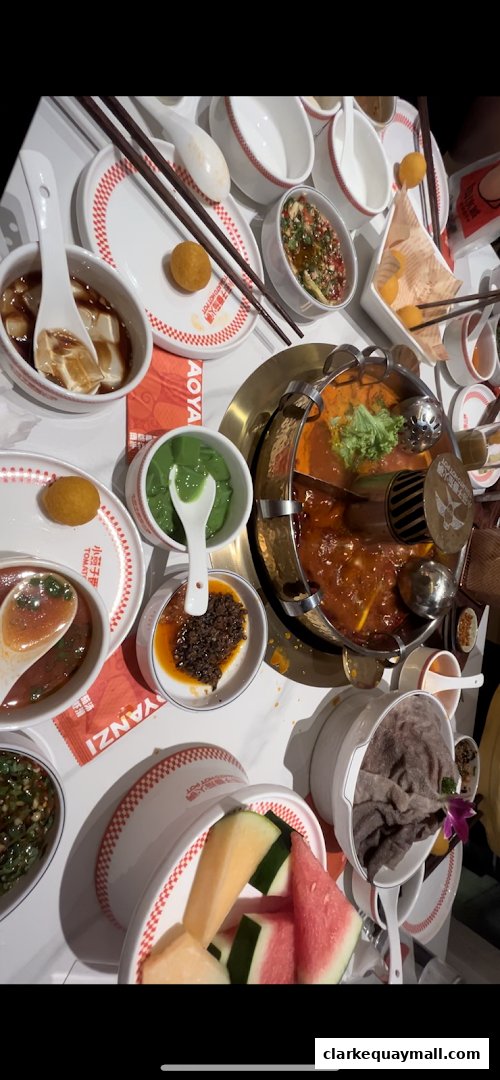 A table laden with various dishes, including a split hot pot, dipping sauces, and fresh fruit.