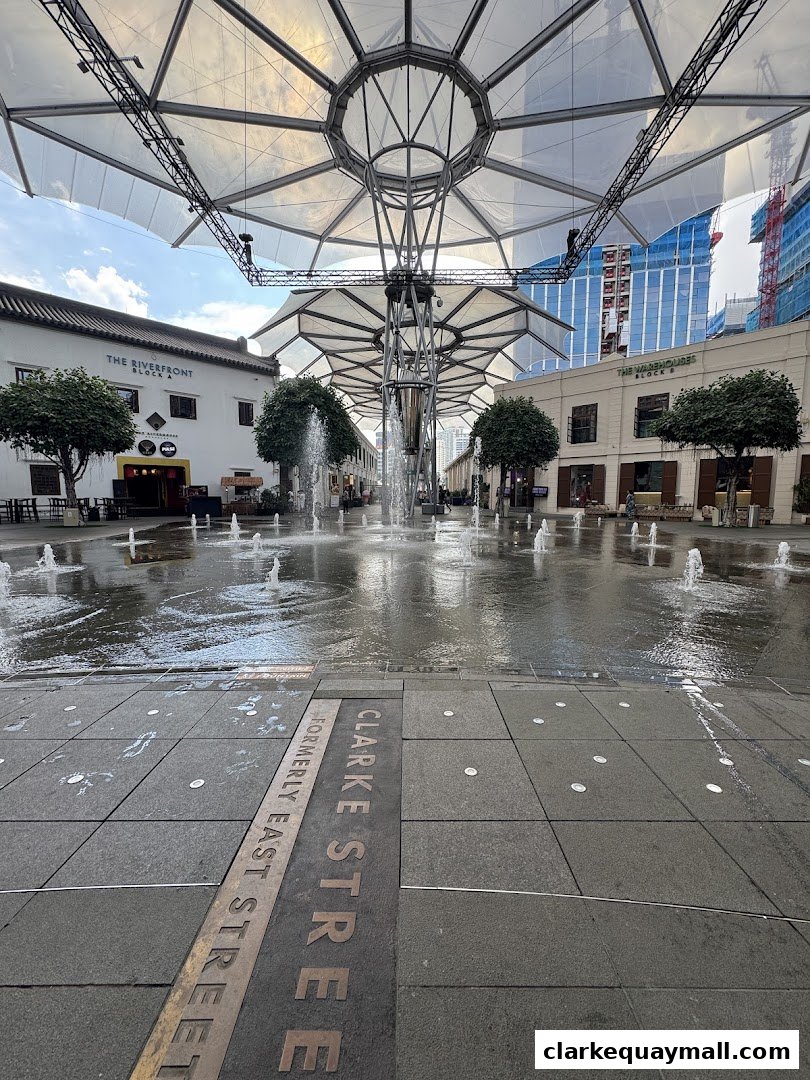 A wide shot of a plaza with fountains and covered walkways, featuring buildings labeled 'The Riverfront' and 'The Warehouses'.