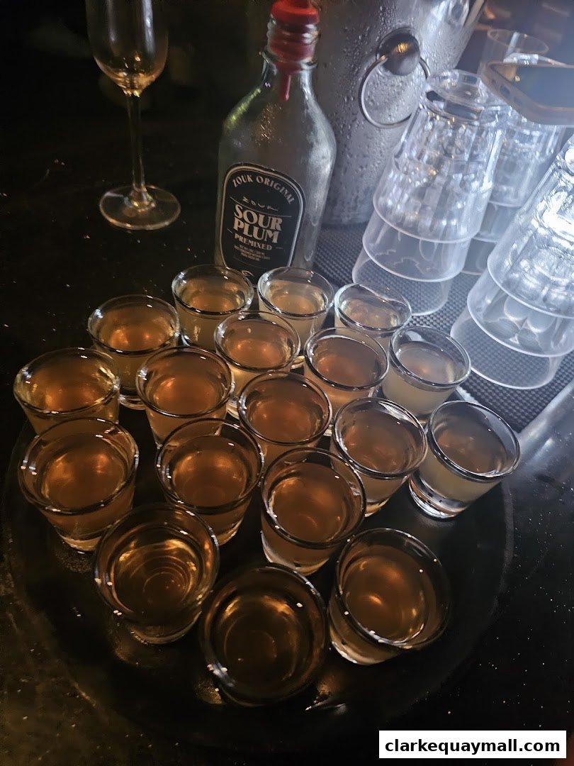 A tray of shot glasses filled with amber liquid next to a bottle of Zouk Sour Plum.