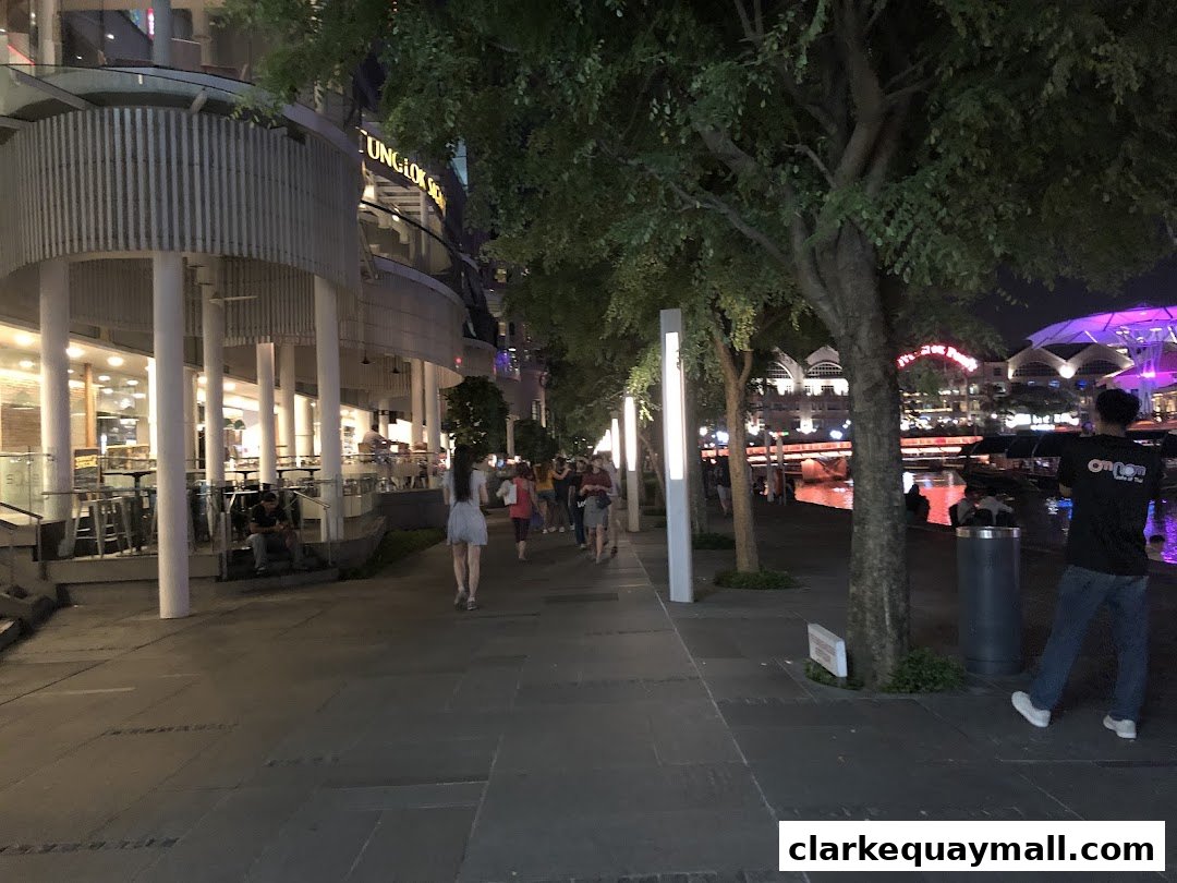 A lively riverside promenade at night with people walking past illuminated shops and trees.
