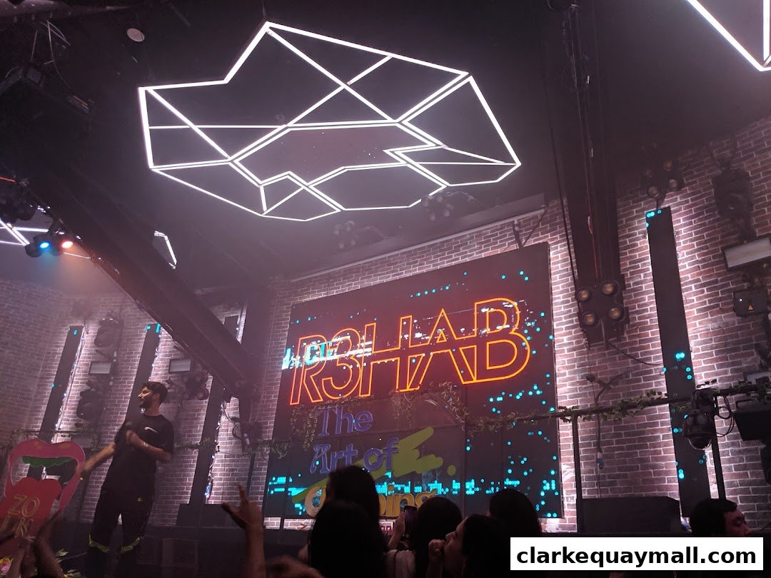 A neon sign reading 'RSHAB The Art of...' illuminates a brick wall in a dimly lit venue.